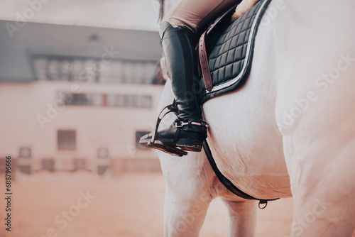 A rider in leather boots sits on a white horse in the saddle. Sports equipment and stirrups. Equestrian sports and horse riding.