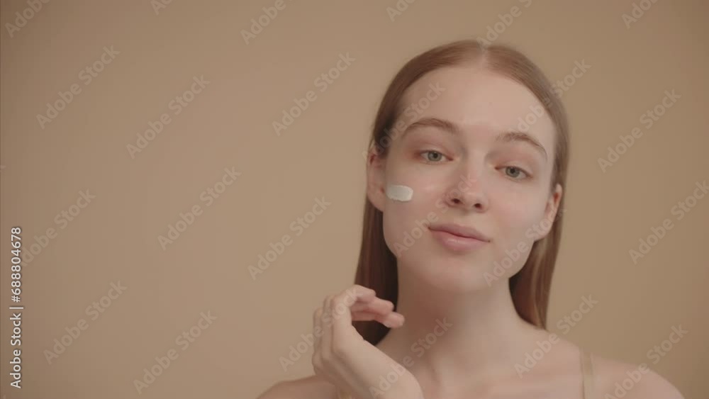 Young lady looking at camera, applying facial cream on cheek. Ad for a facial skin care, anti-aging cosmetic product, facial treatment. Portrait of a young woman on beige background. Slow motion. Young lady looking at camera, applying facial cream on cheek. Ad for a facial skin care, anti-aging cosmetic product, facial treatment. Portrait of a young woman on beige background. Slow motion.