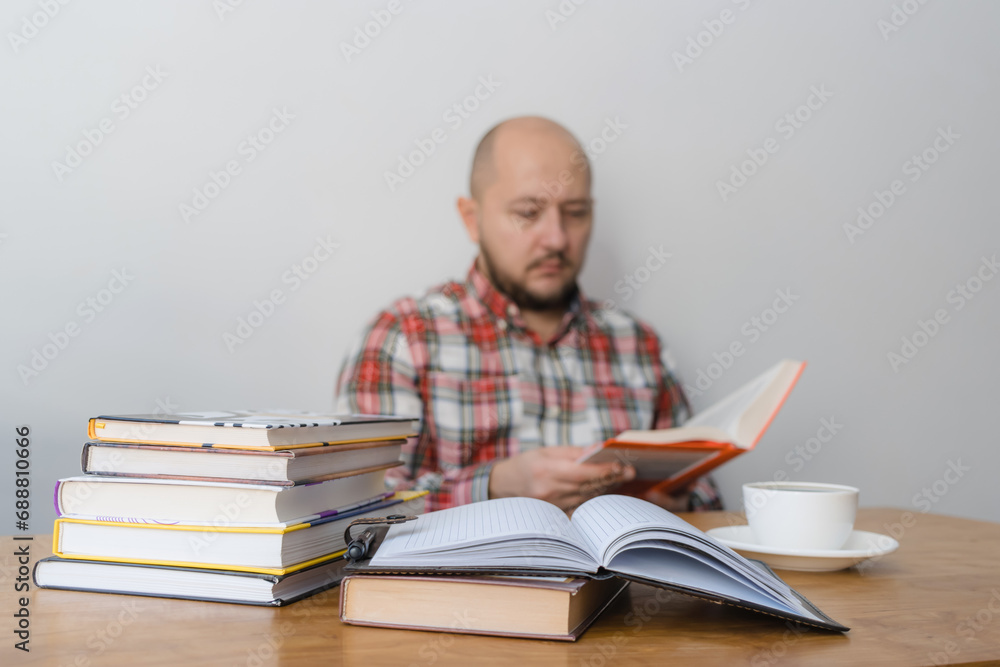 Man reading a book, studying or working, sitting at the table with stack of books and cup of coffee, focus on foreground