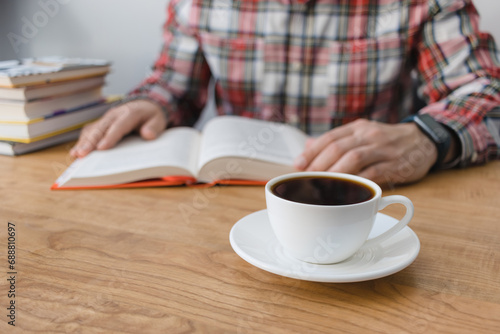 Cup of coffee on wooden table, unrecognizable man reading book, studying or working, sitting at the desk with stack of books, focus on foreground