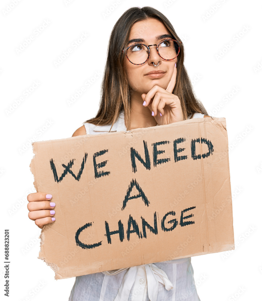 Young hispanic woman holding we need a change banner serious face thinking about question with hand on chin, thoughtful about confusing idea Young hispanic woman holding we need a change banner serious face thinking about question with hand on chin, thoughtful about confusing idea