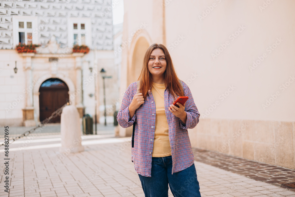 Picture of pretty young woman staying on the street holding phone in hands. 30s tourist in hat walking on old city street checks her smartphone. Use technology concept, Traveling Europe in summer Picture of pretty young woman staying on the street holding phone in hands. 30s tourist in hat walking on old city street checks her smartphone. Use technology concept, Traveling Europe in summer