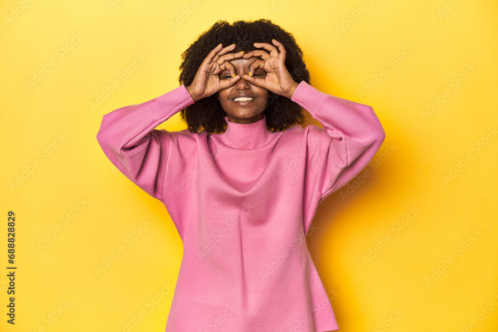 Teen girl in pink sweatshirt, yellow studio backdrop showing okay sign over eyes Teen girl in pink sweatshirt, yellow studio backdrop showing okay sign over eyes