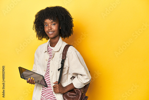 Tech-savvy African-American teen girl with tablet and backpack on yellow studio backdrop.