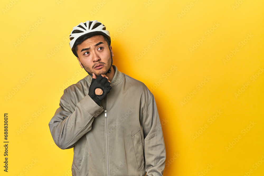 Asian man in bike helmet and gloves, yellow studio backdrop looking sideways with doubtful and skeptical expression. Asian man in bike helmet and gloves, yellow studio backdrop looking sideways with doubtful and skeptical expression.