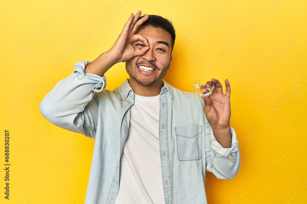 Asian man with invisible dental corrector, yellow studio backdrop excited keeping ok gesture on eye. Asian man with invisible dental corrector, yellow studio backdrop excited keeping ok gesture on eye.