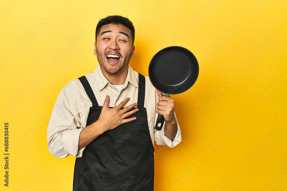 Asian chef holding a pan, yellow studio backdrop laughs out loudly keeping hand on chest. Asian chef holding a pan, yellow studio backdrop laughs out loudly keeping hand on chest.