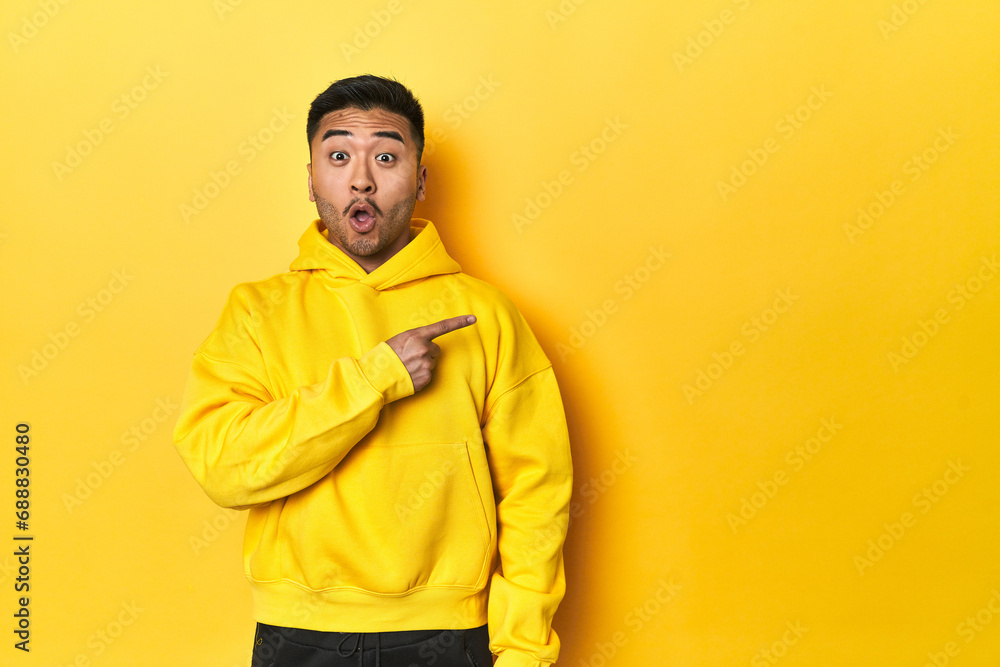 Fototapeta premium Bold Asian man in yellow hoodie on yellow studio pointing to the side