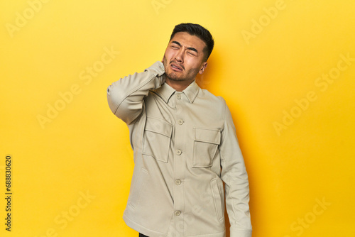 Asian man in stylish gray shirt on yellow studio having a neck pain due to stress, massaging and touching it with hand.