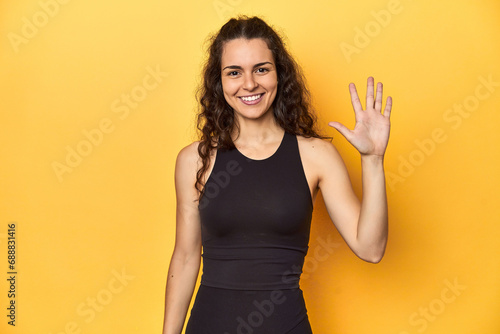 Sporty woman in active wear, yellow backdrop, smiling cheerful showing number five with fingers.