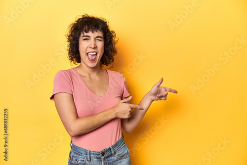 Curly-haired Caucasian woman in pink t-shirt pointing with forefingers to a copy space  expressing excitement and desire.