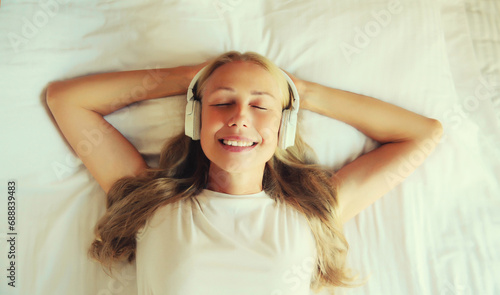Happy relaxed middle-aged woman listening to music with wireless headphones while lying on bed in bedroom at home