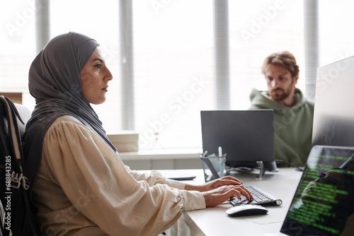 Side view portrait of young Middle-Eastern woman as female programmer using computer in office and wearing headscarf photo