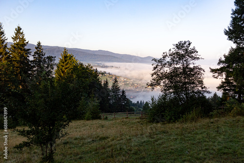 A beautiful fog creeps over the village in the mountains, coniferous and deciduous trees in the foreground