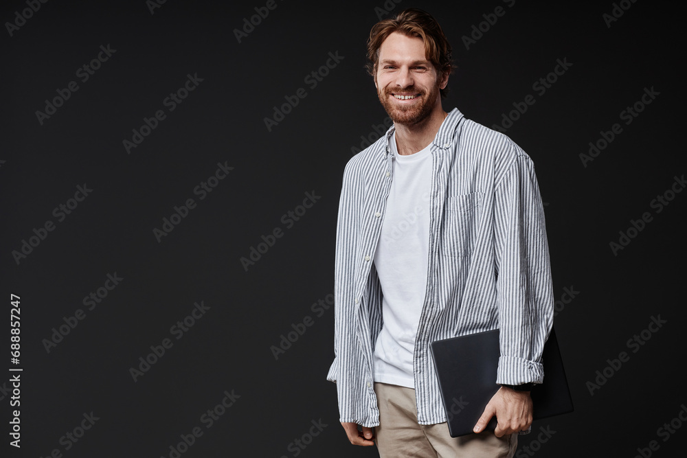 Portrait of smiling bearded man holding laptop and smiling at camera while standing on black background in studio, copy space
