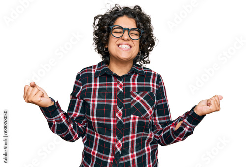 Young hispanic woman with curly hair wearing casual clothes and glasses very happy and excited doing winner gesture with arms raised, smiling and screaming for success. celebration concept.