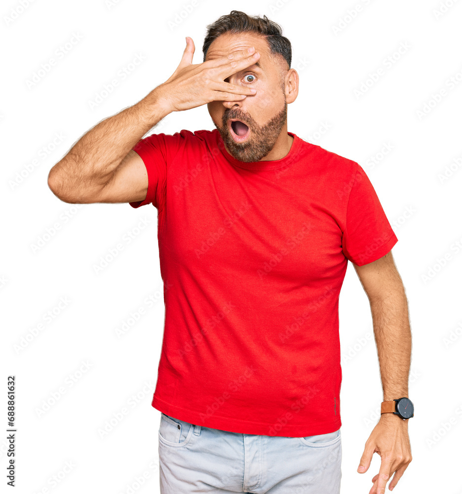 Handsome middle age man wearing casual red tshirt peeking in shock covering face and eyes with hand, looking through fingers with embarrassed expression. Handsome middle age man wearing casual red tshirt peeking in shock covering face and eyes with hand, looking through fingers with embarrassed expression.