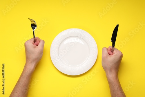 Man holding fork and knife near empty plate at yellow table, top view photo