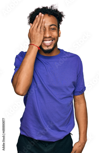 Young african american man with beard wearing casual purple t shirt covering one eye with hand, confident smile on face and surprise emotion.