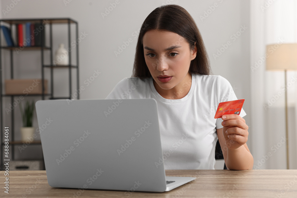 Confused woman with credit card using laptop at table indoors. Be careful - fraud Confused woman with credit card using laptop at table indoors. Be careful - fraud