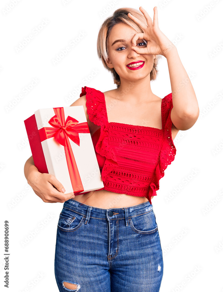 Young beautiful blonde woman holding gift smiling happy doing ok sign with hand on eye looking through fingers Young beautiful blonde woman holding gift smiling happy doing ok sign with hand on eye looking through fingers