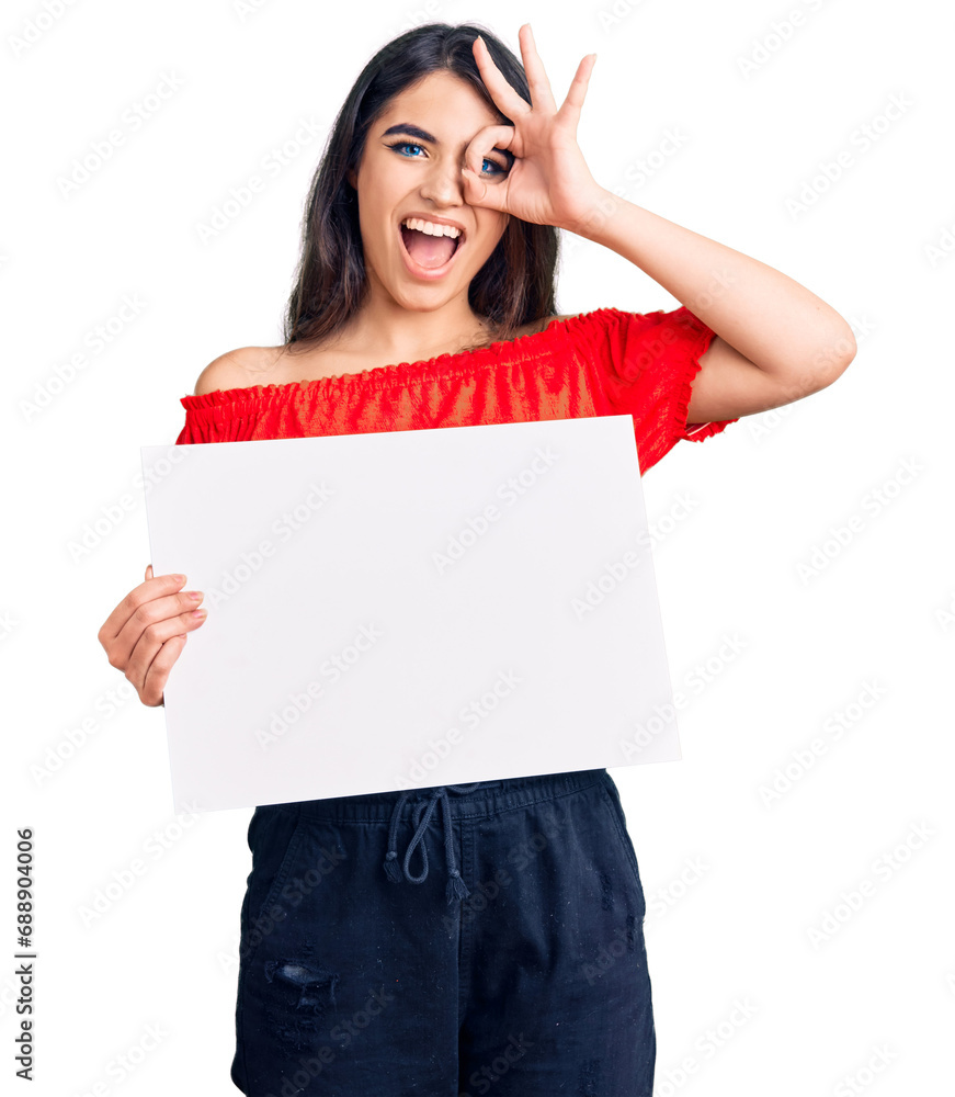 Brunette teenager girl holding blank empty banner smiling happy doing ok sign with hand on eye looking through fingers Brunette teenager girl holding blank empty banner smiling happy doing ok sign with hand on eye looking through fingers