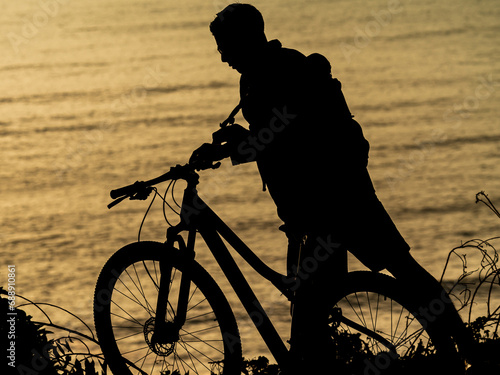 Silueta de ciclista empujando bicicleta en contraluz al atardecer. Determinación palpable mientras el océano se tiñe de tonos cálidos. Fusión de esfuerzo humano y serenidad natural.