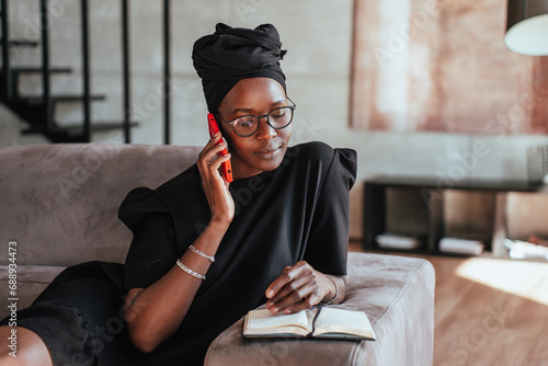 Foto Focused African girl in black turban, glasses sitting on couch talks by phone  looks at opened diary remote working home