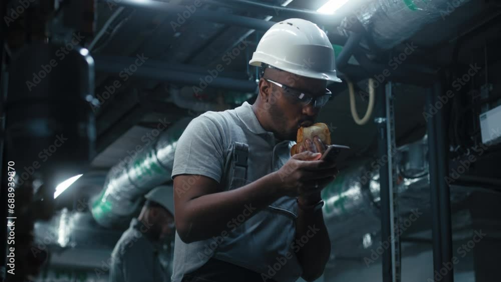 Professional African American engineer in safety uniform, goggles and protective hardhat surfs the Internet using phone and eats sandwich. Industrial worker having break working on modern factory. Professional African American engineer in safety uniform, goggles and protective hardhat surfs the Internet using phone and eats sandwich. Industrial worker having break working on modern factory.