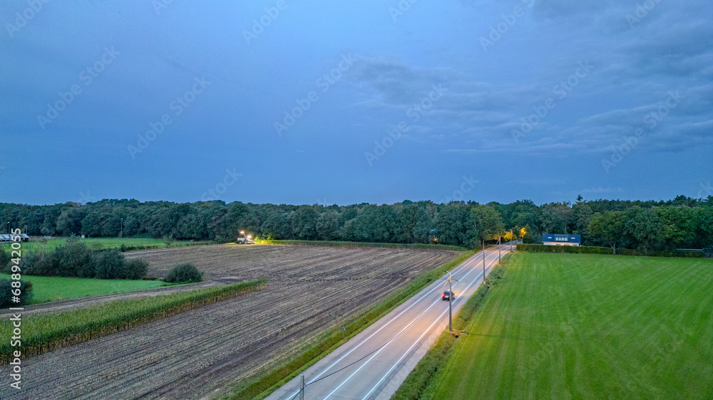 Captured during the tranquil moments of twilight, this image portrays a scenic drive through the countryside. A lone car's headlights cut through the dimming light, illuminating a path along a road