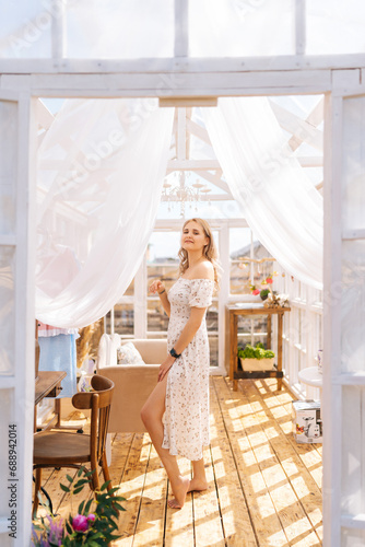 Vertical portrait of charming blonde female in beautiful dress standing inside of summer gazebo house on sunny day, smiling looking at camera with happy expression. Concept of summertime vacation.