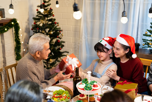 Asian senior grandfather send Christmas gift box and wish grandchild girl all the best while mother embracing and push forward what the thing inside on family dinner party at greeting decoration home