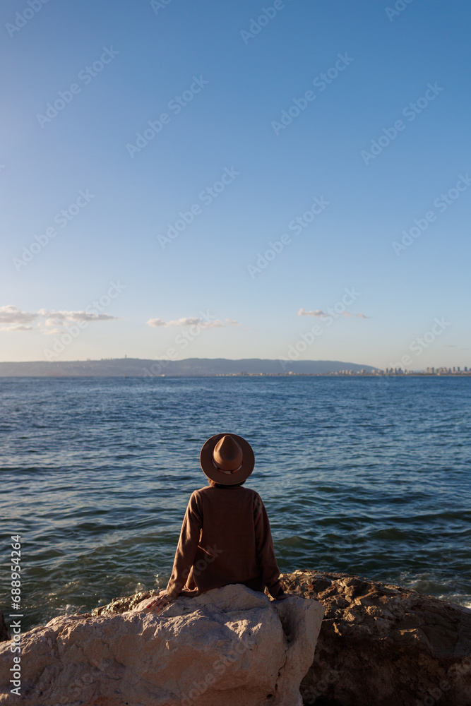 Back view. relaxed girl in a hat and coat sitting in front of the sea on the stones.