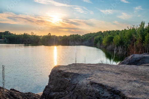 Fototapeta Naklejka Na Ścianę i Meble -  In the soft light of the evening, this image portrays the tranquil end to a day at a rocky lakeshore. The sun, in its final descent, casts a radiant glow that dances across the water's surface. The