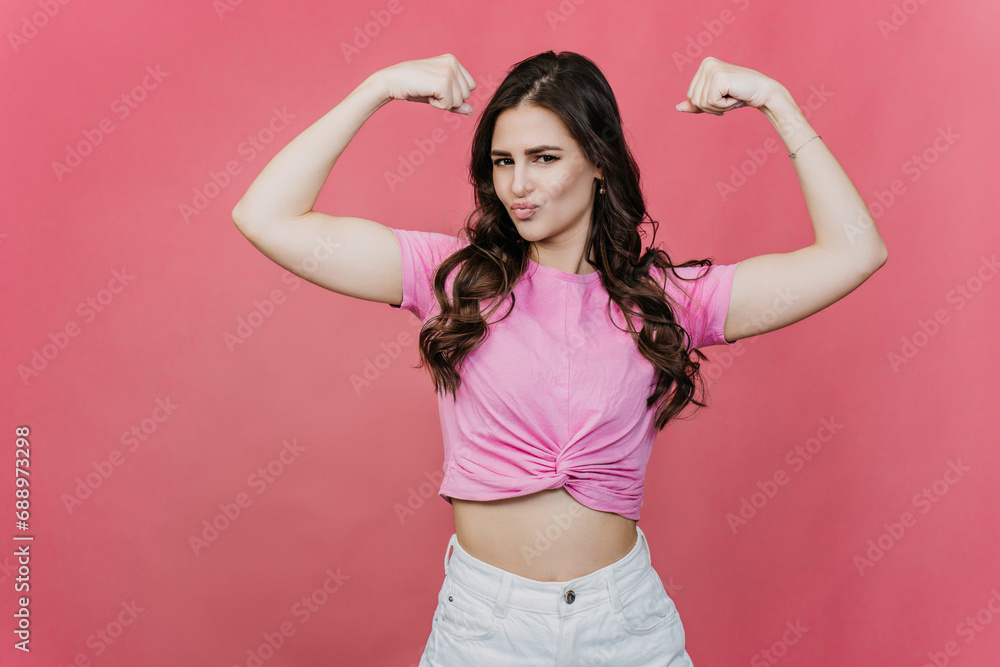 Strong cute brunette girl showing double biceps against studio pink background looks at camera, radiates confidence. Purposeful people. Strength concept. Strong cute brunette girl showing double biceps against studio pink background looks at camera, radiates confidence. Purposeful people. Strength concept.