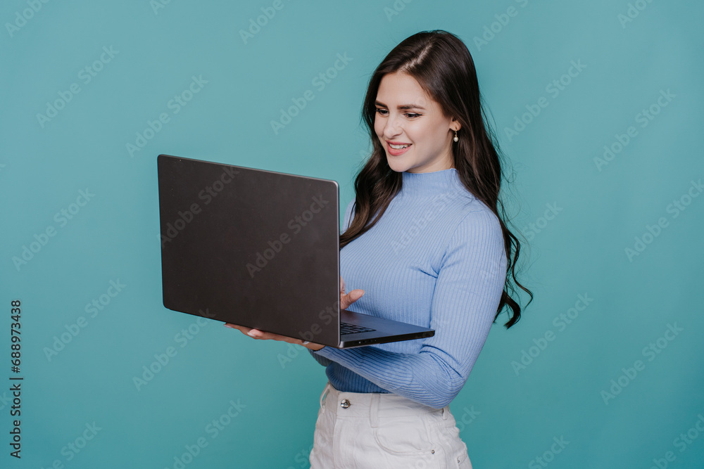 Joyful brunette girl in casual clothe holds laptop standing against turquoise background toothy smiles working. Remote work. Young woman using computer. Joyful brunette girl in casual clothe holds laptop standing against turquoise background toothy smiles working. Remote work. Young woman using computer.