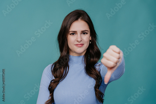 Brunette caucasian girl with wavy hair looks at camera shows thumb down gesture, negative decision standing against turquoise background. Don’t do this, its not good choice. photo
