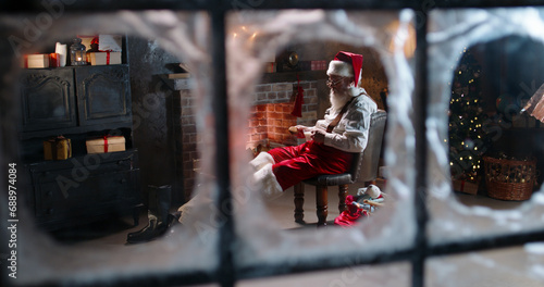 Tired Santa Claus sleeping near fireplace, holding cookies and cup with milk outside view through window. Overworked Mr Claus relaxing, sitting with legs outstretched. House of Santa, free time.