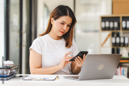 Cheerful Asian woman confidently uses mobile phone, tablet with laptop and notebooks on the desk in a bright office.