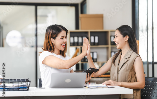 Two Asian colleagues, freelance writer and graphic designer discussing about creating characters in screenplays work and smiling with tablets and a laptop, journalist, artist, giving high five
