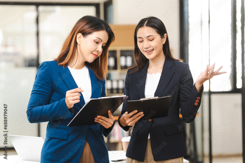 Two middle aged and young Asian female executives in formal suits review bar chart, discussing business strategies in office setting, senior executives or directors in advertising or public relations Two middle aged and young Asian female executives in formal suits review bar chart, discussing business strategies in office setting, senior executives or directors in advertising or public relations