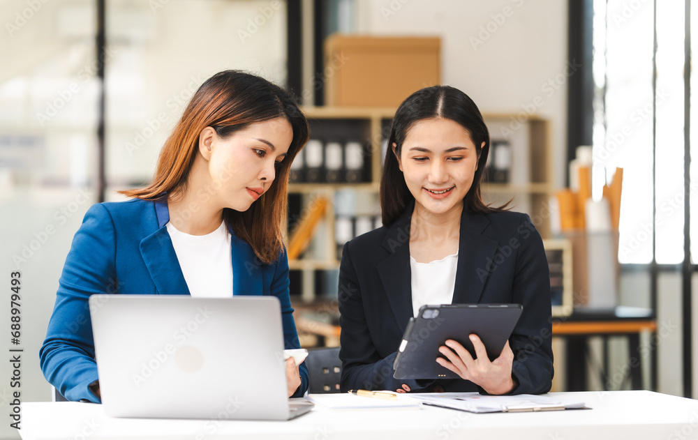 Two middle aged and young Asian female executives in formal suits review bar chart, discussing business strategies in office setting, senior executives or directors in advertising or public relations