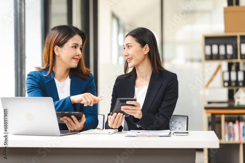 Two middle aged and young Asian female executives in formal suits review bar chart, discussing business strategies in office setting, senior executives or directors in advertising or public relations