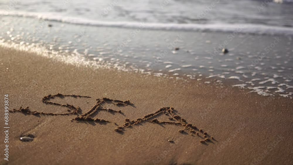 SEA figures hand written in the golden sand on beautiful sunset golden light. The sea wave washes away the inscription SEA on the yellow sand close-up. SEA figures hand written in the golden sand on beautiful sunset golden light. The sea wave washes away the inscription SEA on the yellow sand close-up.