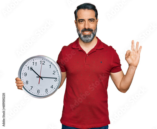 Middle aged man with beard holding big clock doing ok sign with fingers, smiling friendly gesturing excellent symbol