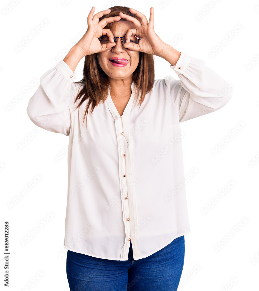 Middle age latin woman wearing casual clothes and glasses doing ok gesture like binoculars sticking tongue out, eyes looking through fingers. crazy expression.