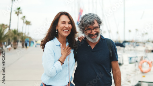 Elderly couple in love are happy on walk together, stand embracing in the port and talk on yacht background