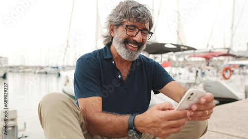 Konstfotografi Happy senior man in glasses sits in the port on yacht background uses smartphone