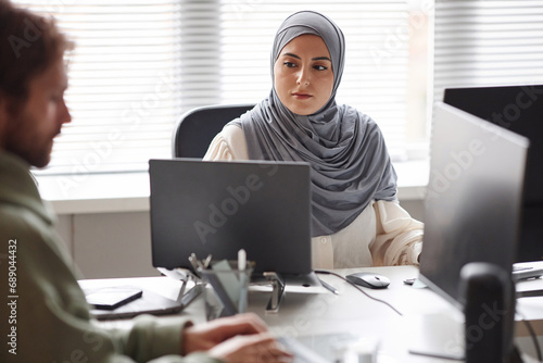 Front view portrait of young Middle Eastern woman looking at laptop screen while working with multiple computer devices in office