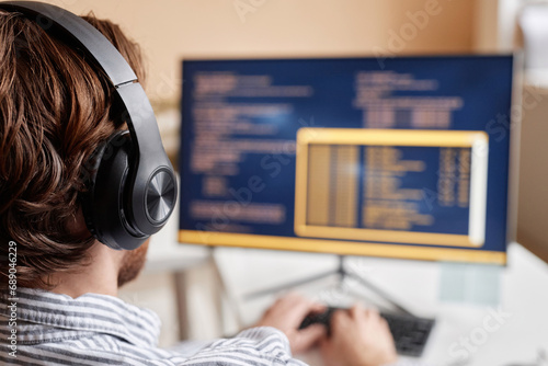 Over shoulder of male computer programmer writing code in office and wearing big headphones, copy space